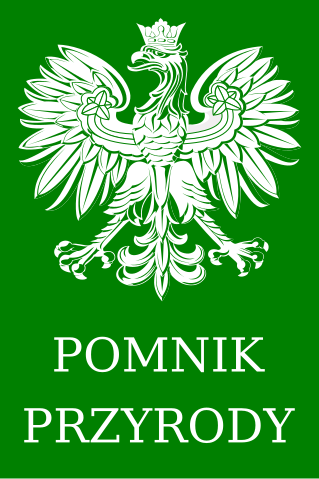Tabliczka urzędowa Pomnik przyrody. Na zielonym tle przedstawiono w białym kolorze na górze godło państwowe a poniżej napis drukowanymi literami w dwóch rzędach: POMNIK PRZYRODY