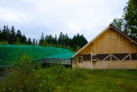 Widok na wolierę pokazową głuszców w Jaworzynce Wyrchczadeczce (gmina Istebna) - po prawej stronie drewniany budynek z trójkątnym dachem, połączony z wybiegiem po lewej stronie, pokrytym od góry zieloną siatką, w tle las.