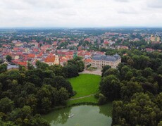 Widok z góry na park pszczyński z pałacem oraz w tle zabudowania starego miasta, otoczone gęstą zielenią i małym stawem z łódką na pierwszym planie pod zachmurzonym niebem.