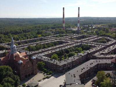 Widok z góry (zdjęcie z drona) na zabytkowy układ zabudowań w dzielnicy Nikiszowiec, na pierwszym planie po lewej strony widok na neobarokowy kościół św. Anny, w oddali za Nikiszem  dwa kominy Kopalni Wieczorek.