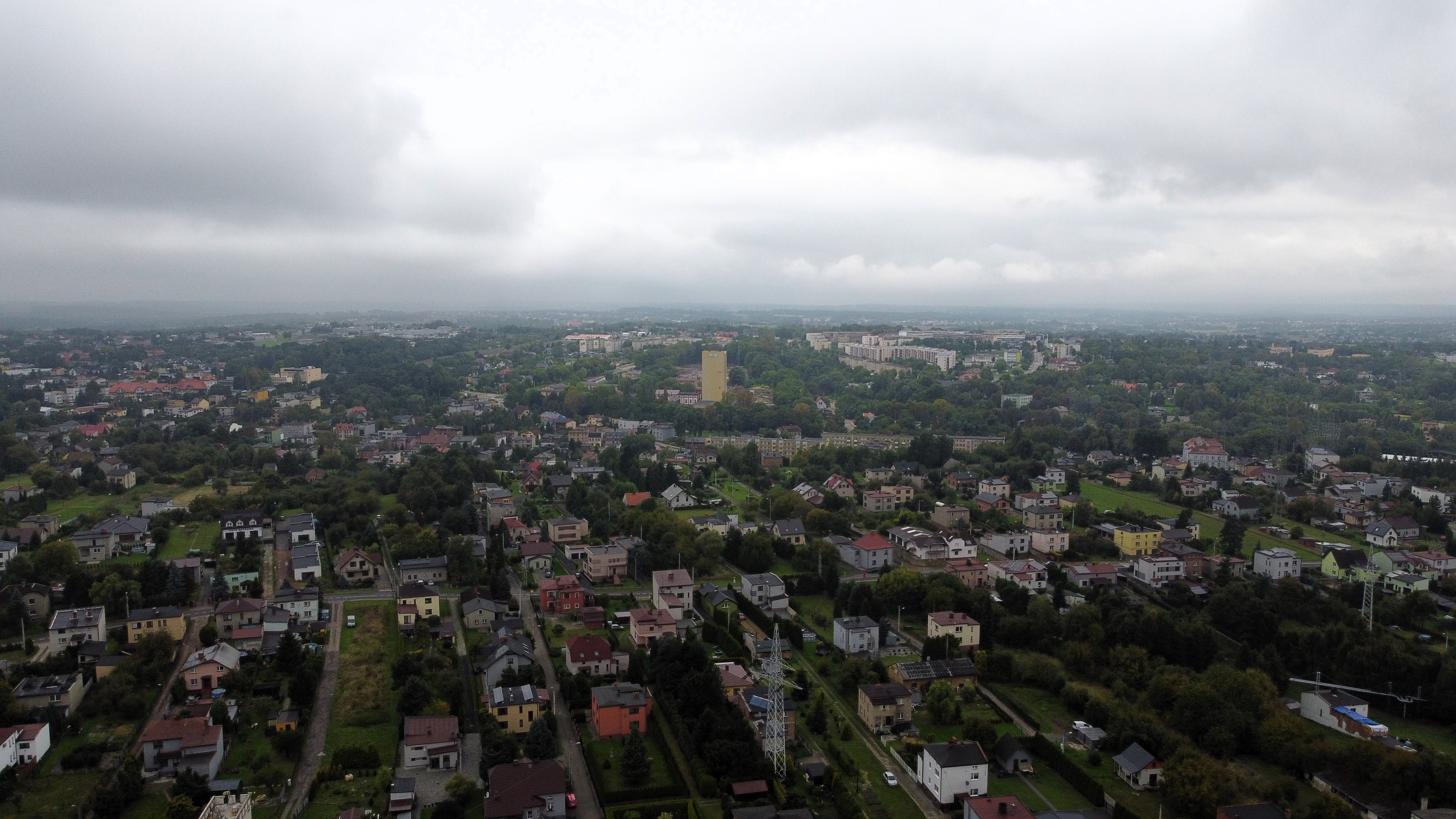 Widok z góry na miasto o charakterze współczesnym, rozproszona, zróżnicowana zabudowa, pomiędzy zabudowaniami zieleń miejska, na górze szare, zachmurzone niebo.