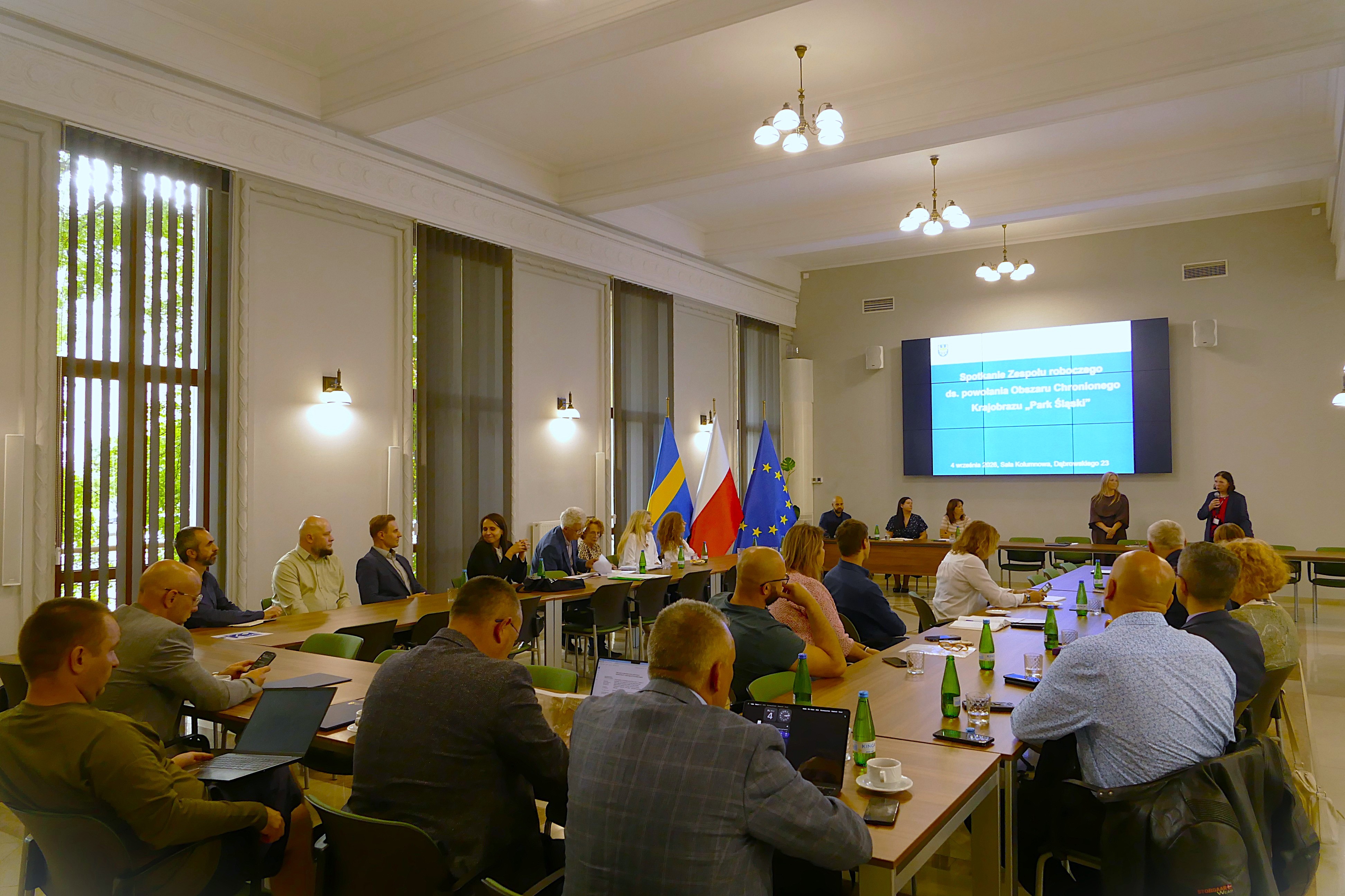 Spotkanie zespołu roboczego w sali konferencyjnej. Uczestnicy siedzą przy stołach, a na ekranie wyświetlana jest prezentacja dotycząca powołania obszaru chronionego krajobrazu „Park Śląski”. W spotkaniu uczestniczy kilkadziesiąt osób, część korzysta z laptopów, a przy stole prezydialnym prowadzone są wystąpienia i dyskusja. Salę oświetlają wiszące i boczne lampy, a wysokie okna wpuszczają światło dzienne.