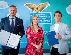 Zdjęcie przedstawia trzy osoby podpisujące umowę na realizację II edycji kampanii edukacyjno-informacyjnej „Bierzemy powietrze pod swoje skrzydła”, w tle plakat promocyjny kampanii.