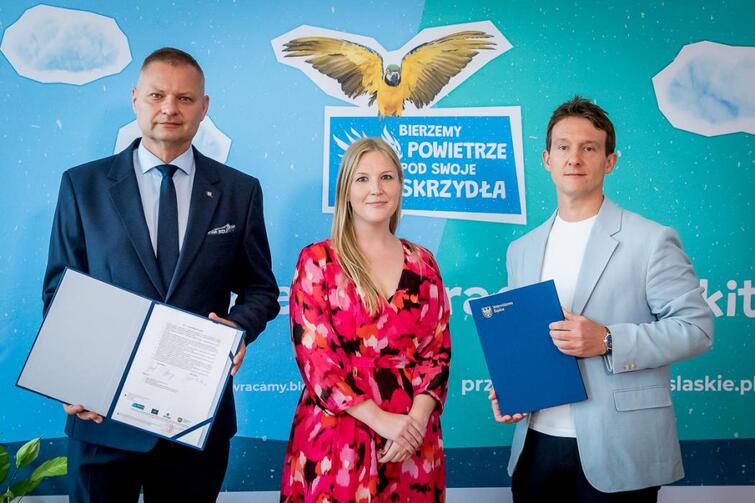 Zdjęcie przedstawia trzy osoby podpisujące umowę na realizację II edycji kampanii edukacyjno-informacyjnej „Bierzemy powietrze pod swoje skrzydła”, w tle plakat promocyjny kampanii.