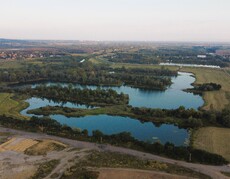 Widok z góry na zbiornik wodny rozdzielony groblami z krzewami, dookoła tereny zielone, w oddali zarys zabudowy miejskiej/wiejskiej.