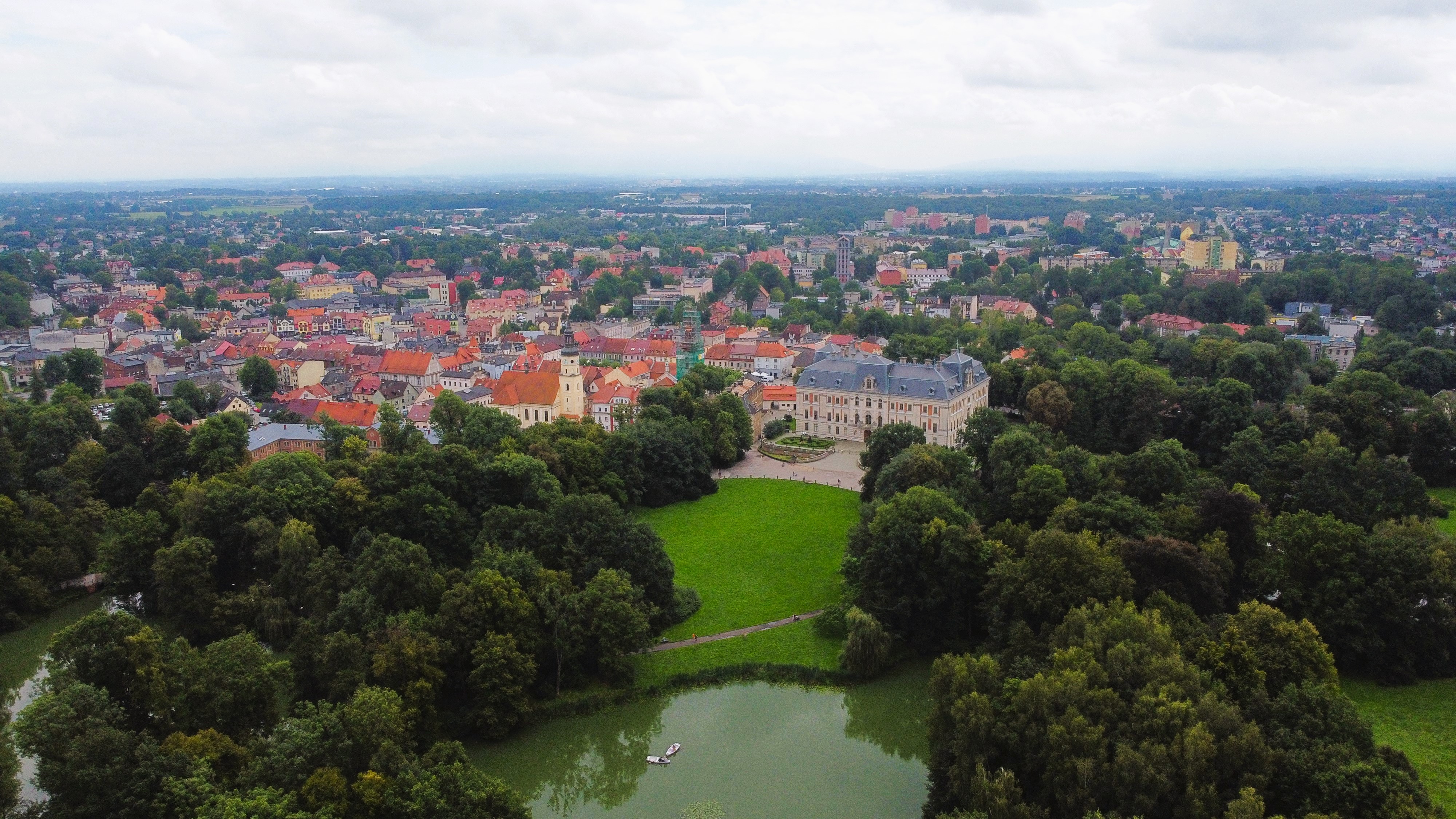 Widok z góry na park pszczyński z pałacem oraz w tle zabudowania starego miasta, otoczone gęstą zielenią i małym stawem z łódką na pierwszym planie pod zachmurzonym niebem.